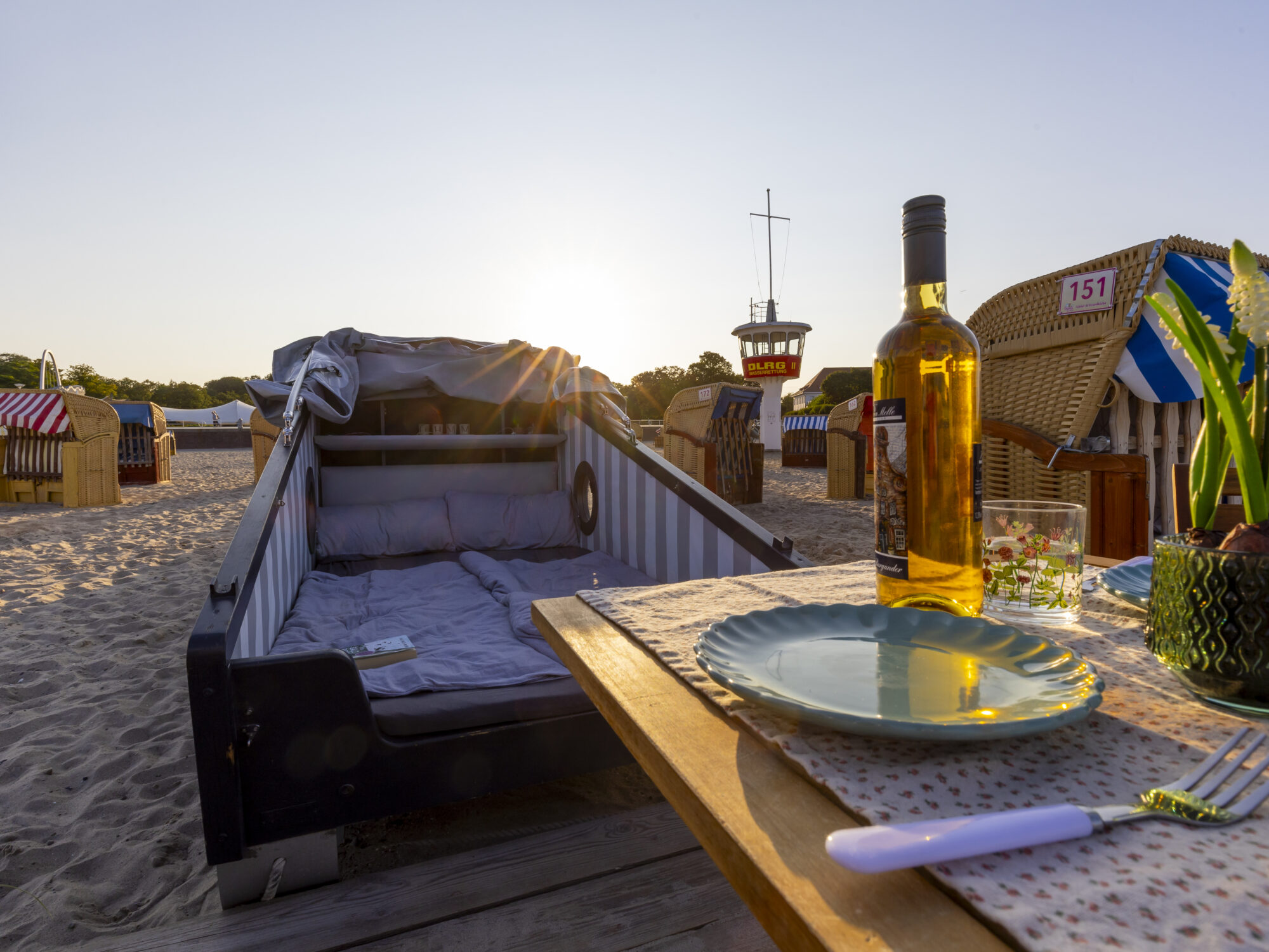 7 × Sommer in Lübeck erleben 20 Ein gedeckter Tisch mit einer Flasche Wein und Geschirr steht neben einem geöffneten Schlafstrandkorb am Strand. Im Hintergrund sind weitere Strandkörbe und ein Rettungsturm zu sehen, während die Sonne untergeht.
